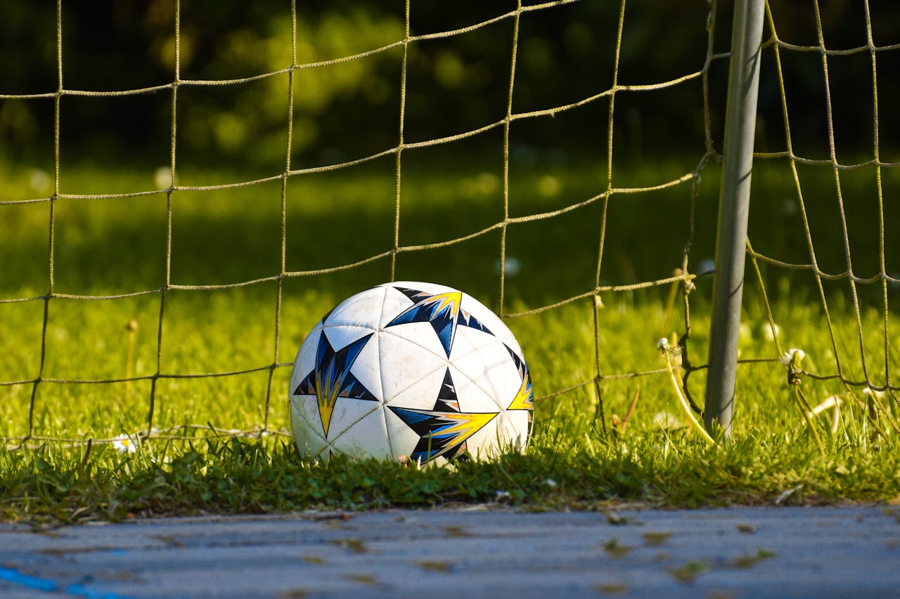Close-up of a soccer ball resting on a grass field against the goal net in natural daylight.